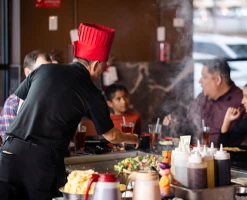 A chef wearing a red hat cooks food on a hibachi grill in front of seated customers at a restaurant.