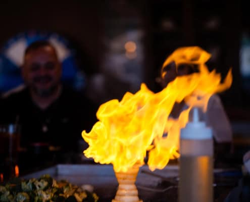 Large yellow flame rises from a grill at a dining table, with blurred people and a condiment bottle visible in the background.