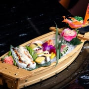 A wooden boat platter filled with assorted sushi, sashimi, and garnishes, including flowers and a decorative plastic dragon, on a black marble table.