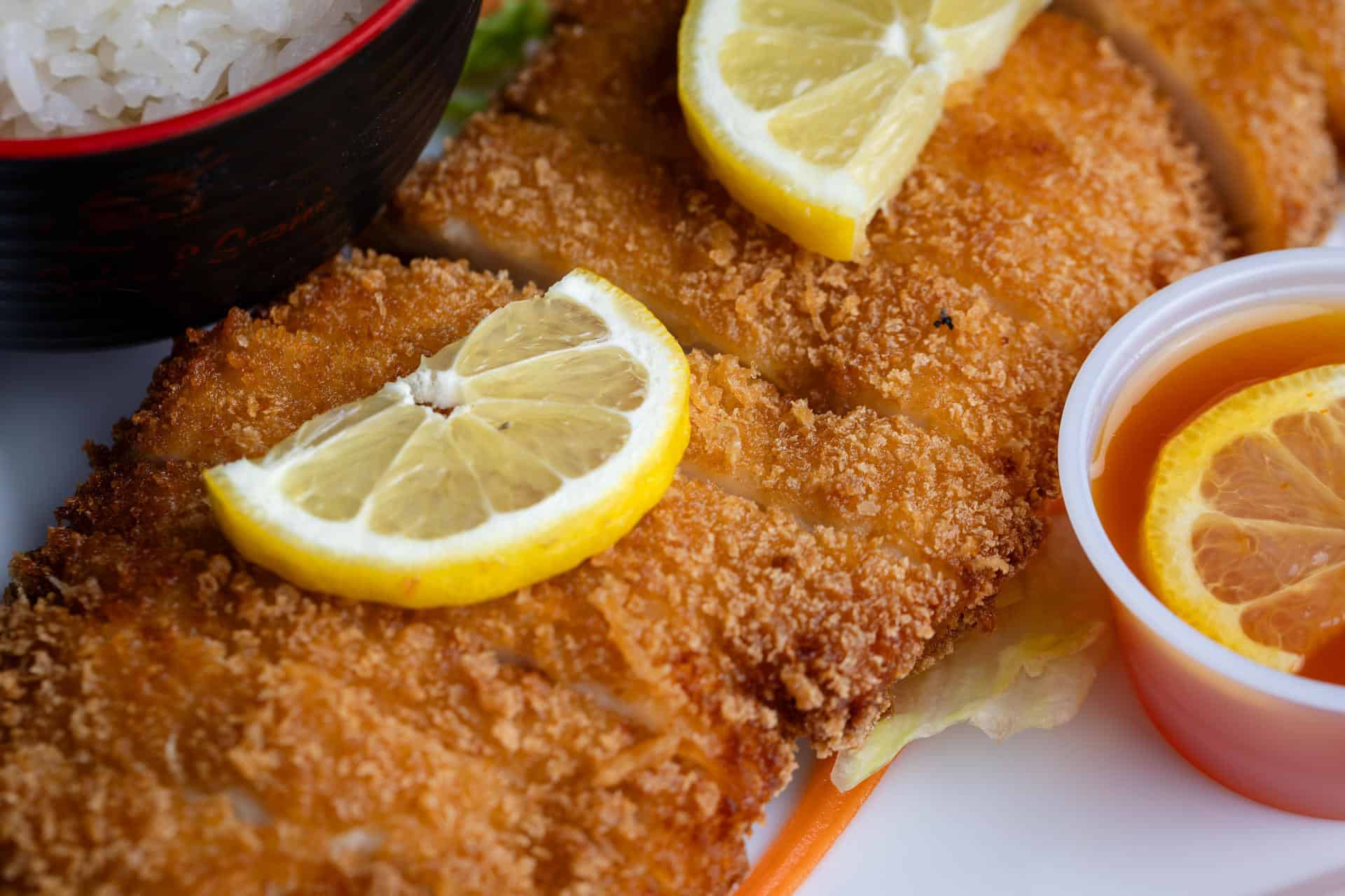 Close-up of breaded fried cutlet topped with lemon slices, served with a side of rice in a bowl and a small container of orange dipping sauce with a lemon slice.